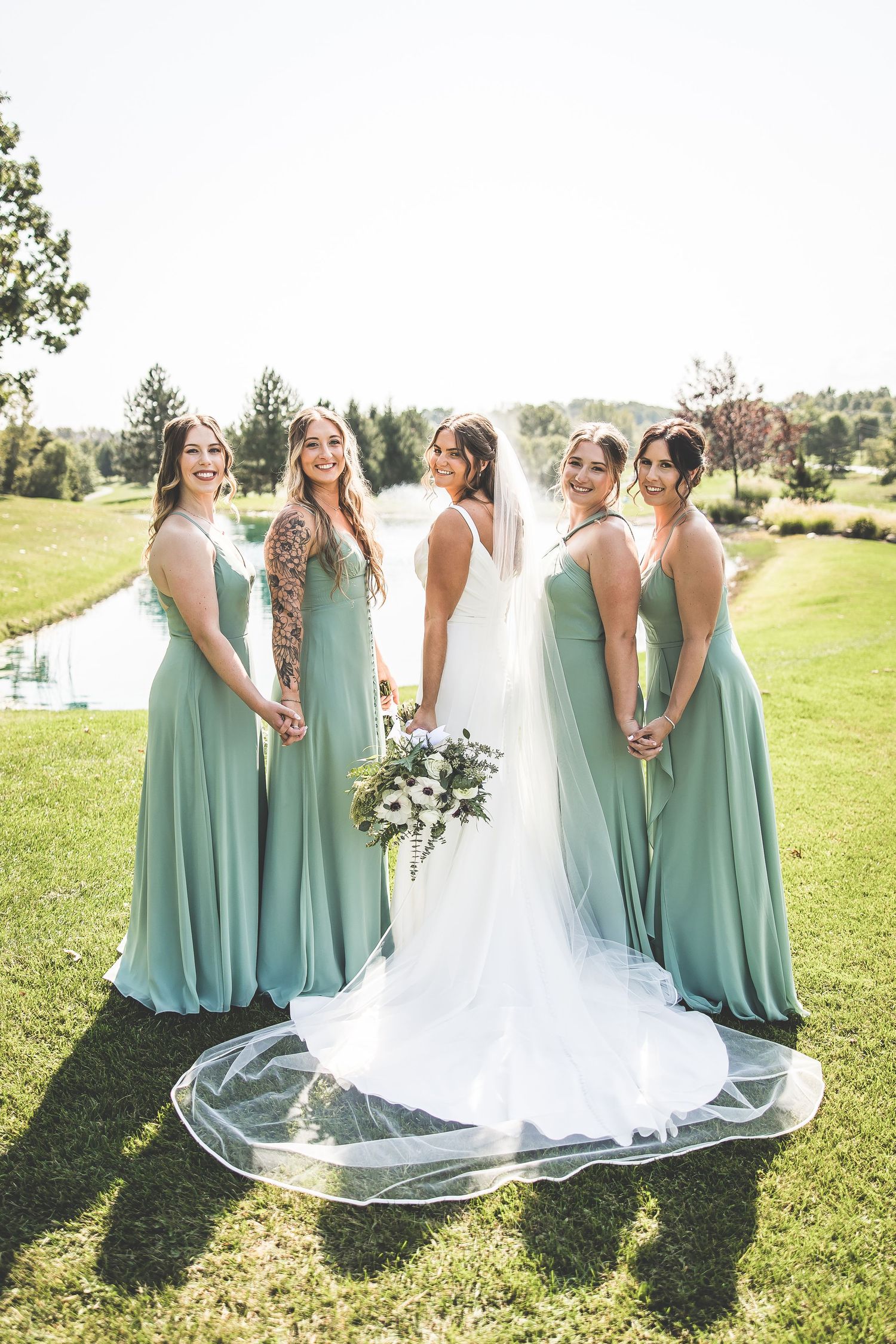 Bridesmaids in mint green dresses stand together with bride in flowing white gown on sunny lawn.