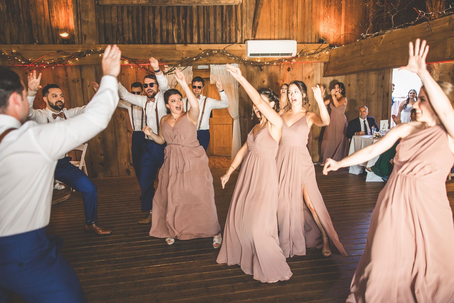 Wedding reception dance party in rustic barn venue with string lights and pink bridesmaid dresses.