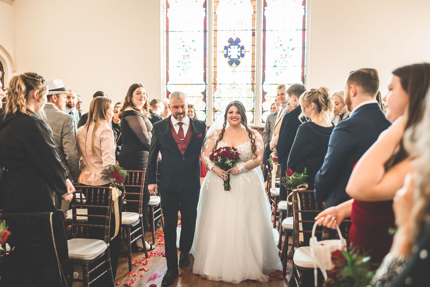 Wedding ceremony guests seated in church pews with stained glass windows.