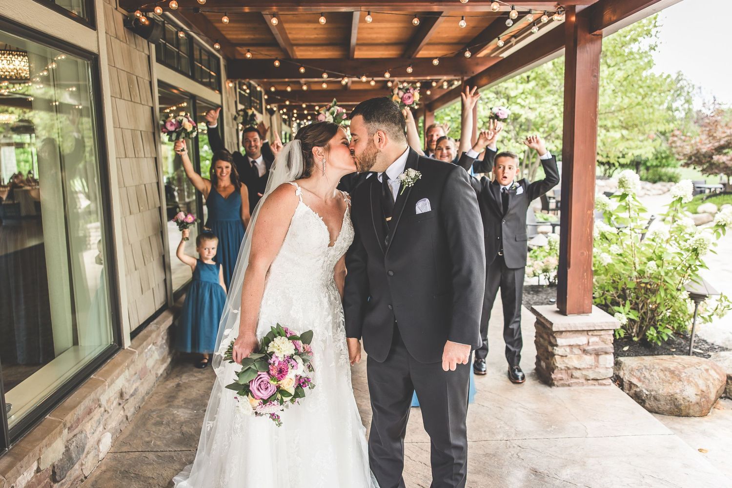 Wedding party gathers under covered outdoor venue with string lights and stone details.