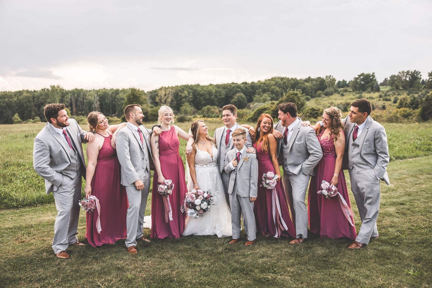 Panoramic wedding party photo with bridesmaids in pink and groomsmen in gray suits on grassy hillside.