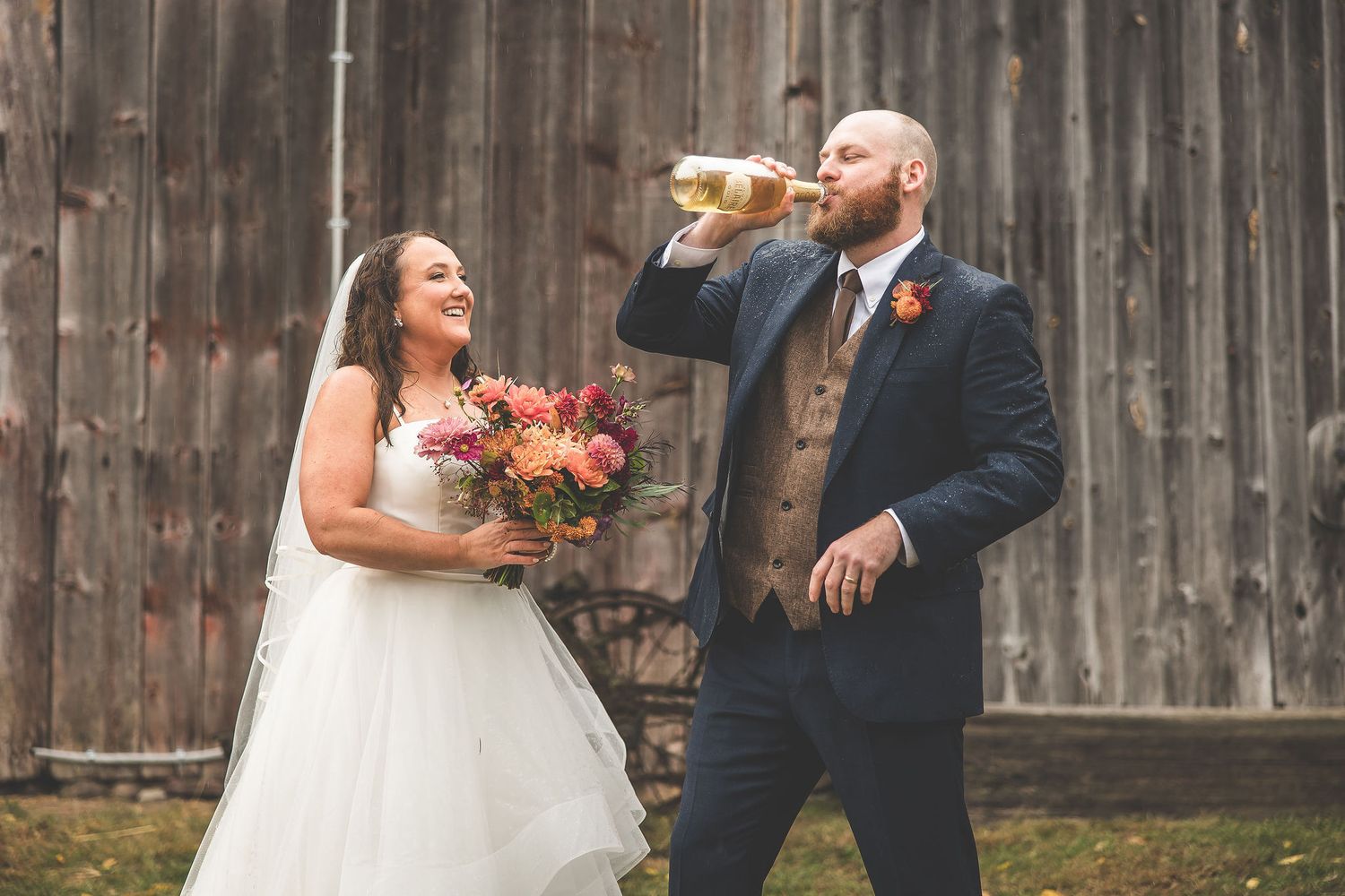 Newlyweds share a joyful moment with autumn-colored bouquet against weathered barn wood.