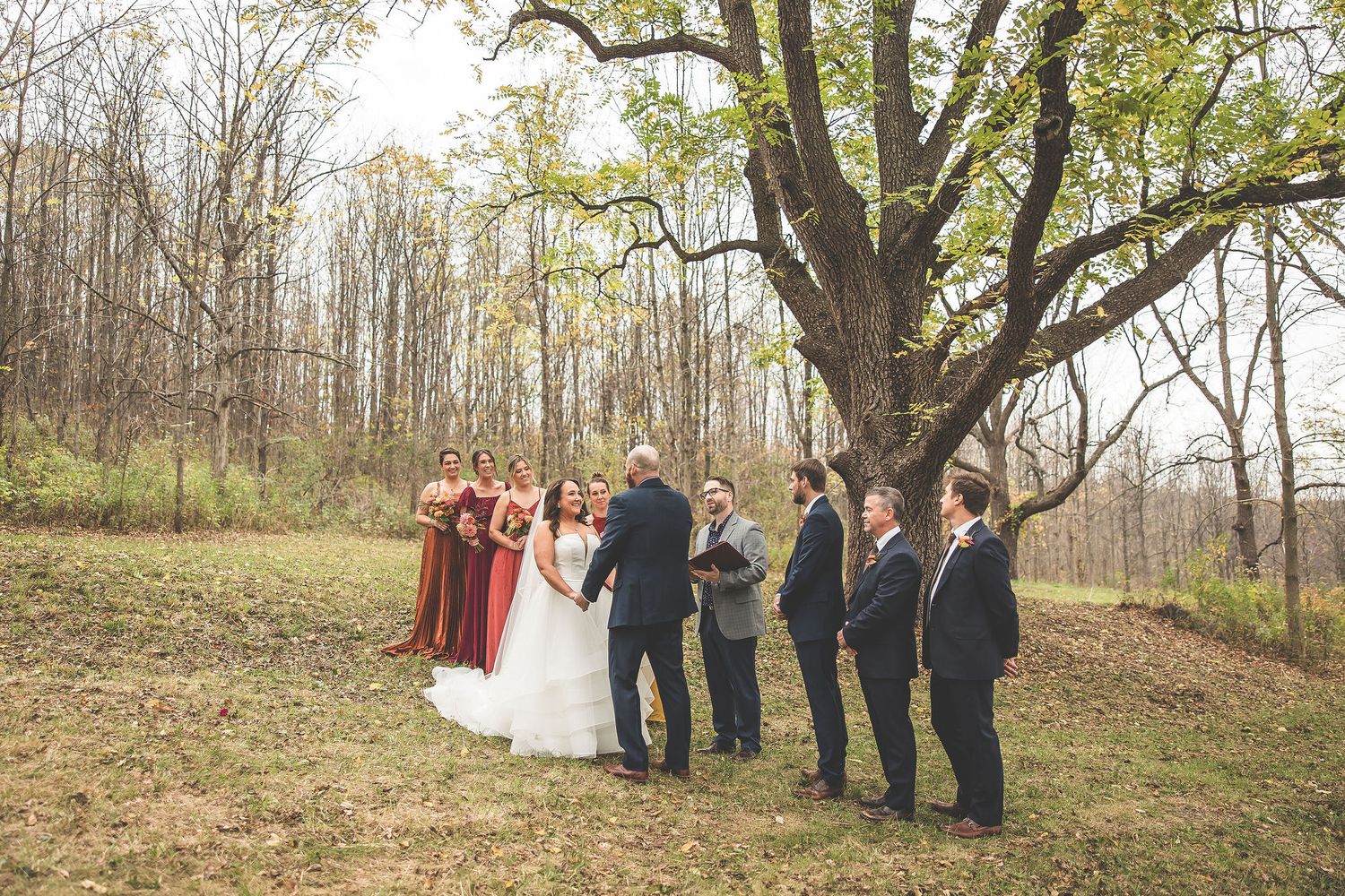 Wedding ceremony taking place on hillside under bare autumn trees.