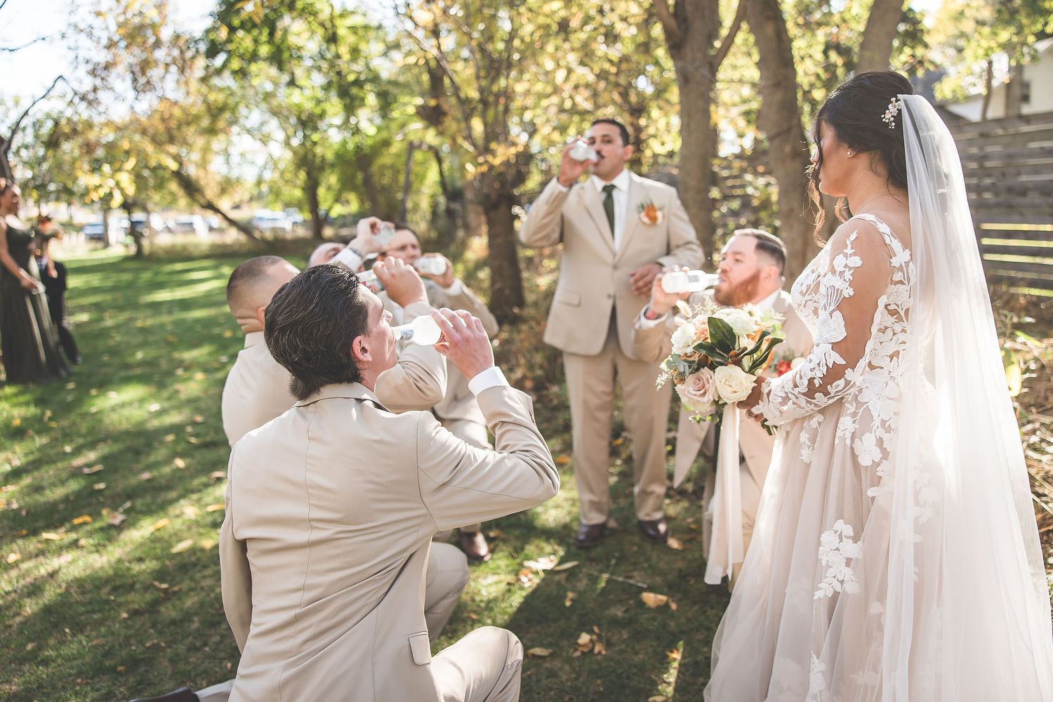Wedding celebration scene with guests in light colored formal attire in a sunlit outdoor garden.