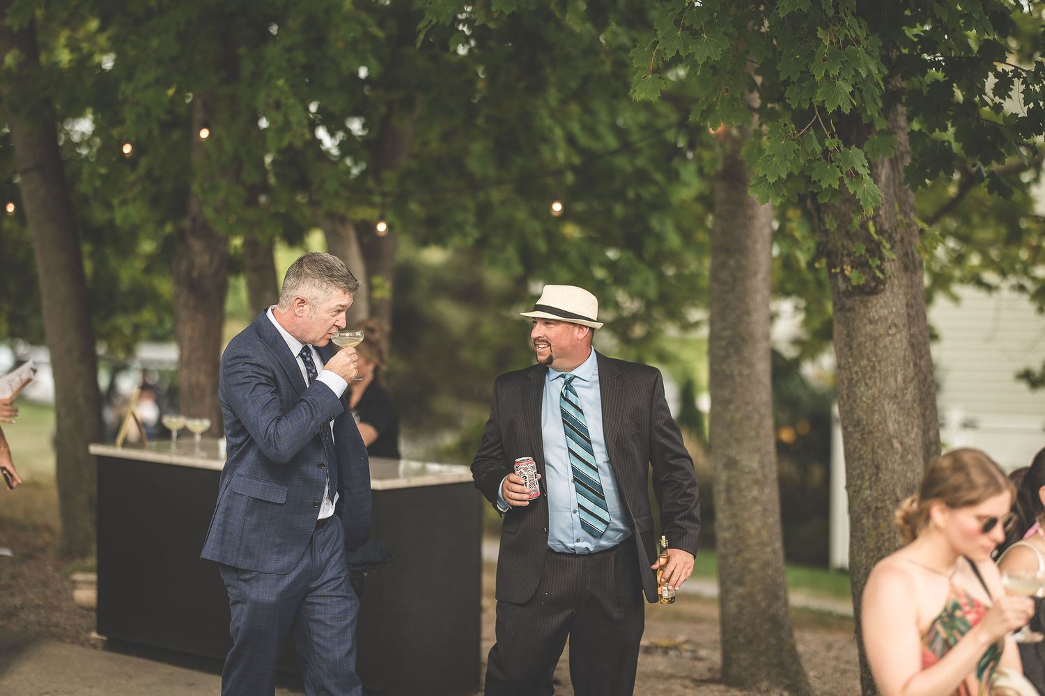Wedding guests mingle during an outdoor cocktail hour beneath tall trees.