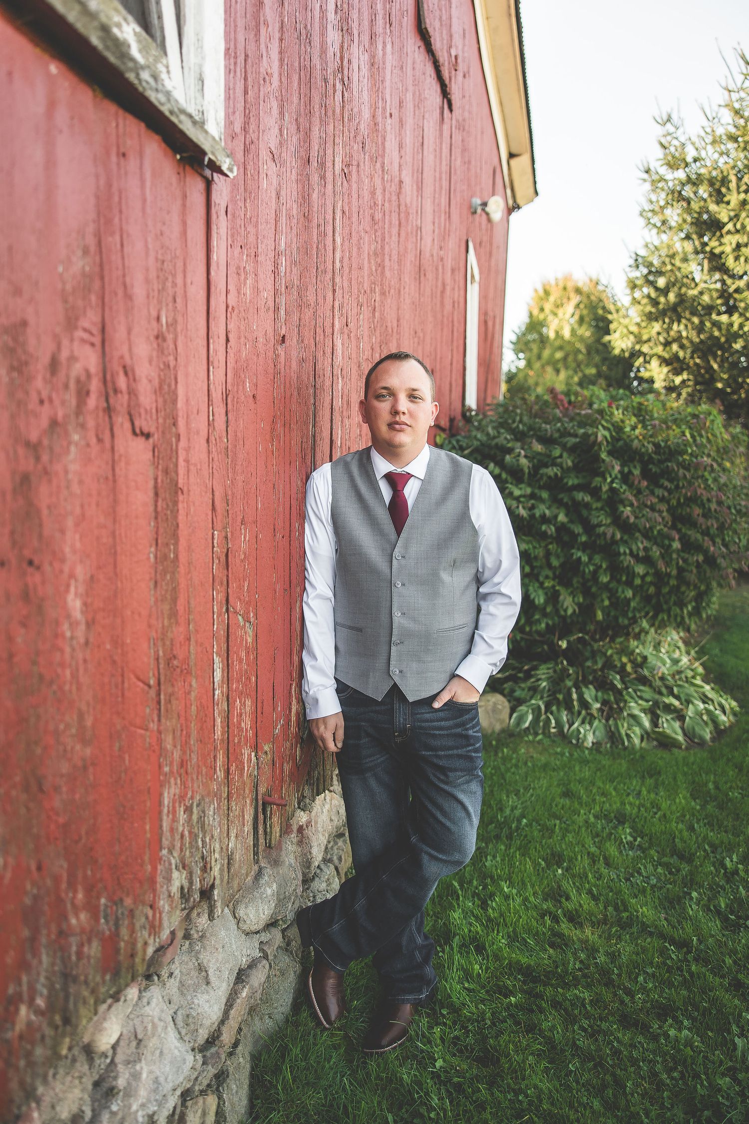 Groom in gray vest poses against rustic red barn wall in outdoor wedding photos.