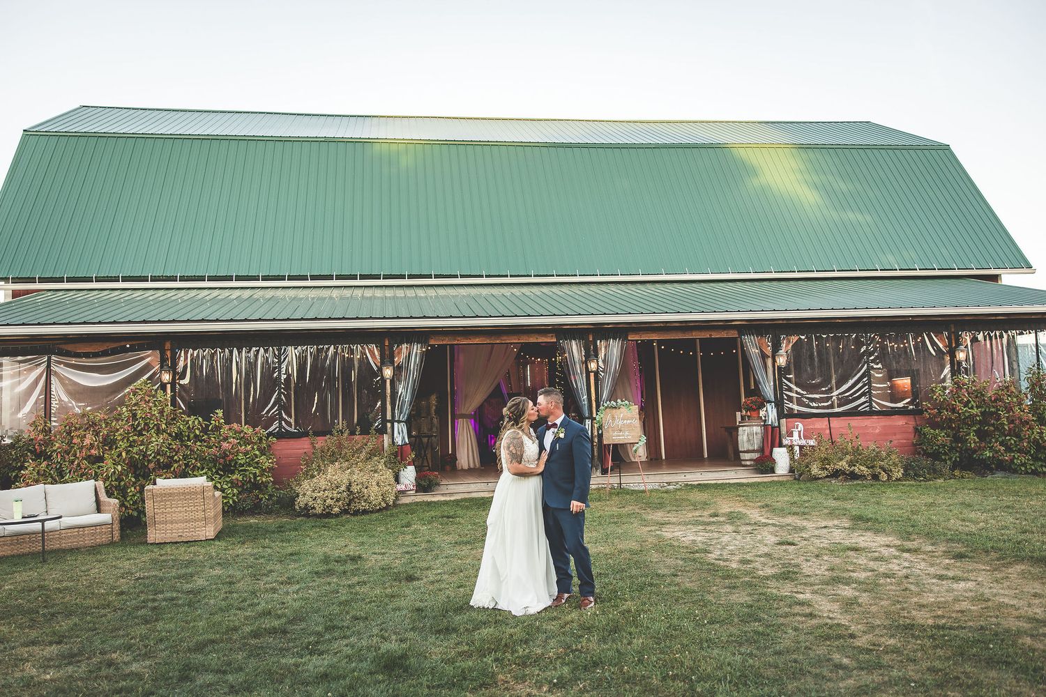 Wedding couple stands in front of green-roofed rustic barn venue.