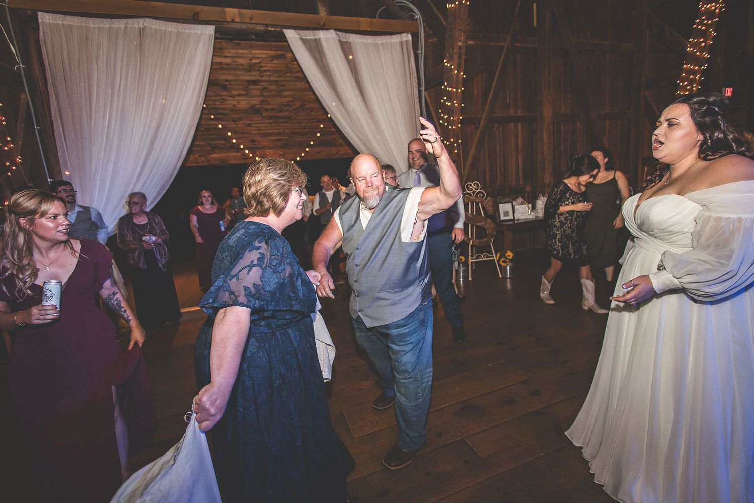 Wedding guests dance joyfully in a rustic barn venue decorated with flowing white drapes and string lights.