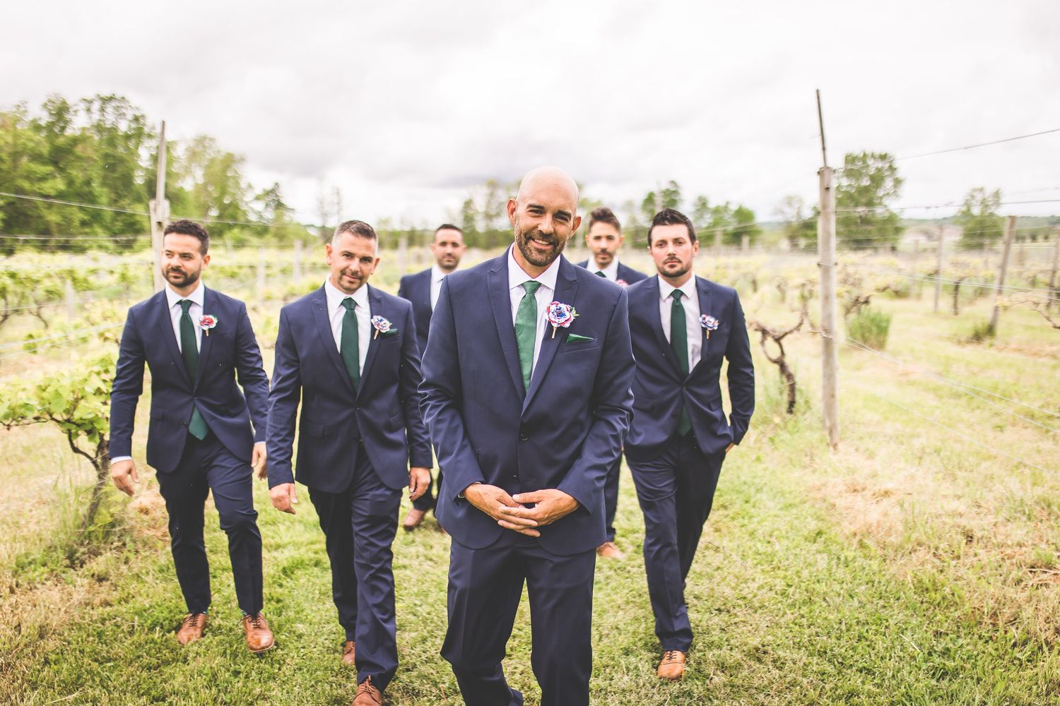 Wedding party walks through vineyard with groomsmen in navy suits.