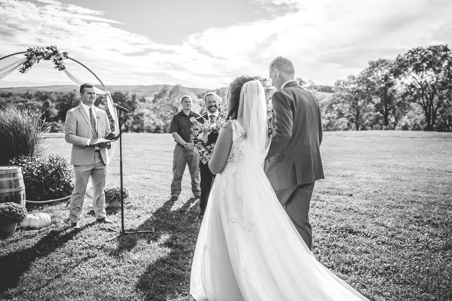 Black and white photo of a wedding ceremony taking place outdoors with rolling hills in the background.