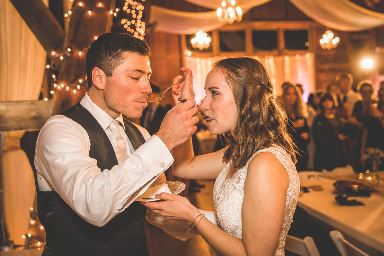 Wedding guests dance in a warmly lit rustic venue with string lights and decorative draping.