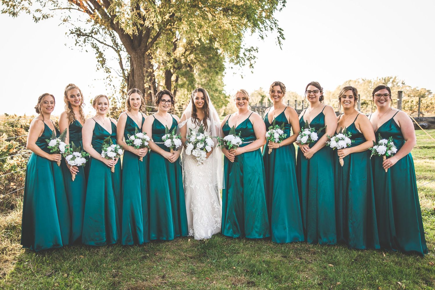 Large wedding party in teal bridesmaid dresses and suits poses together on a sunny hillside.
