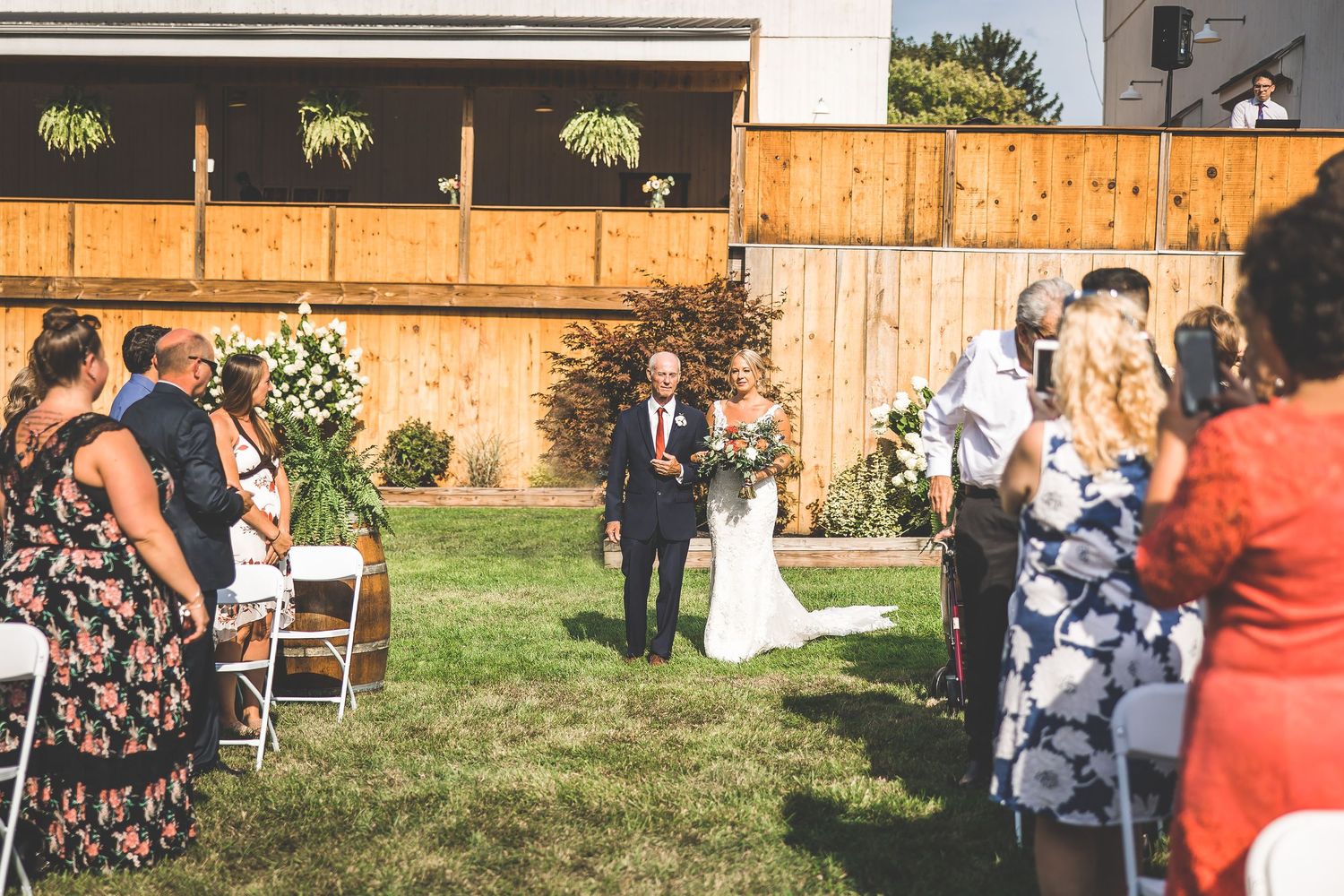 Outdoor wedding ceremony with guests seated on white chairs near rustic barn.