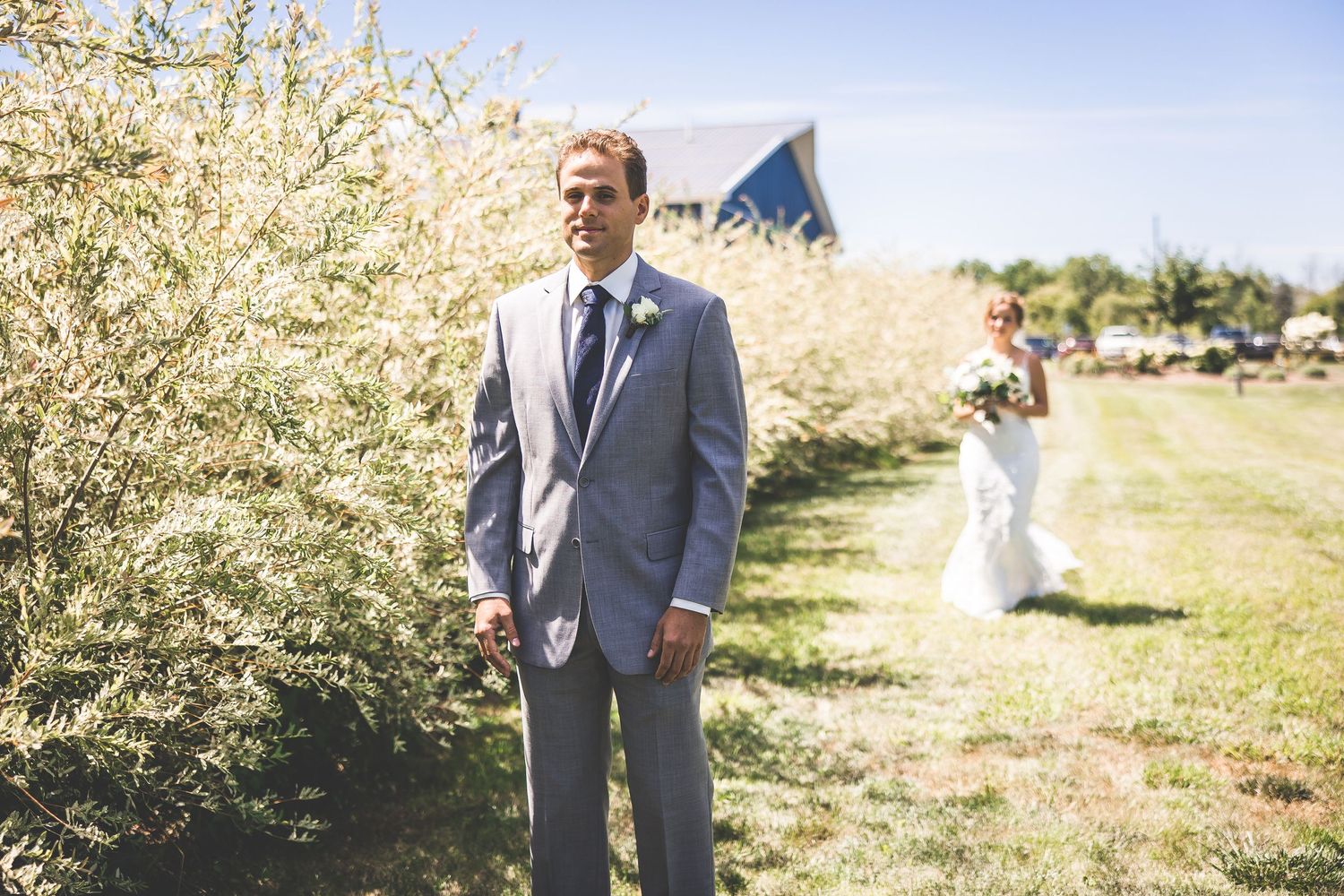 Wedding couple poses in front of blooming orchard trees on a sunny day.