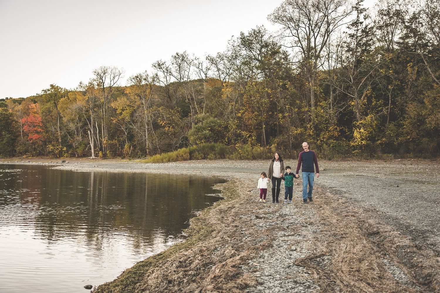 People walking along rocky shoreline with autumn trees in background.