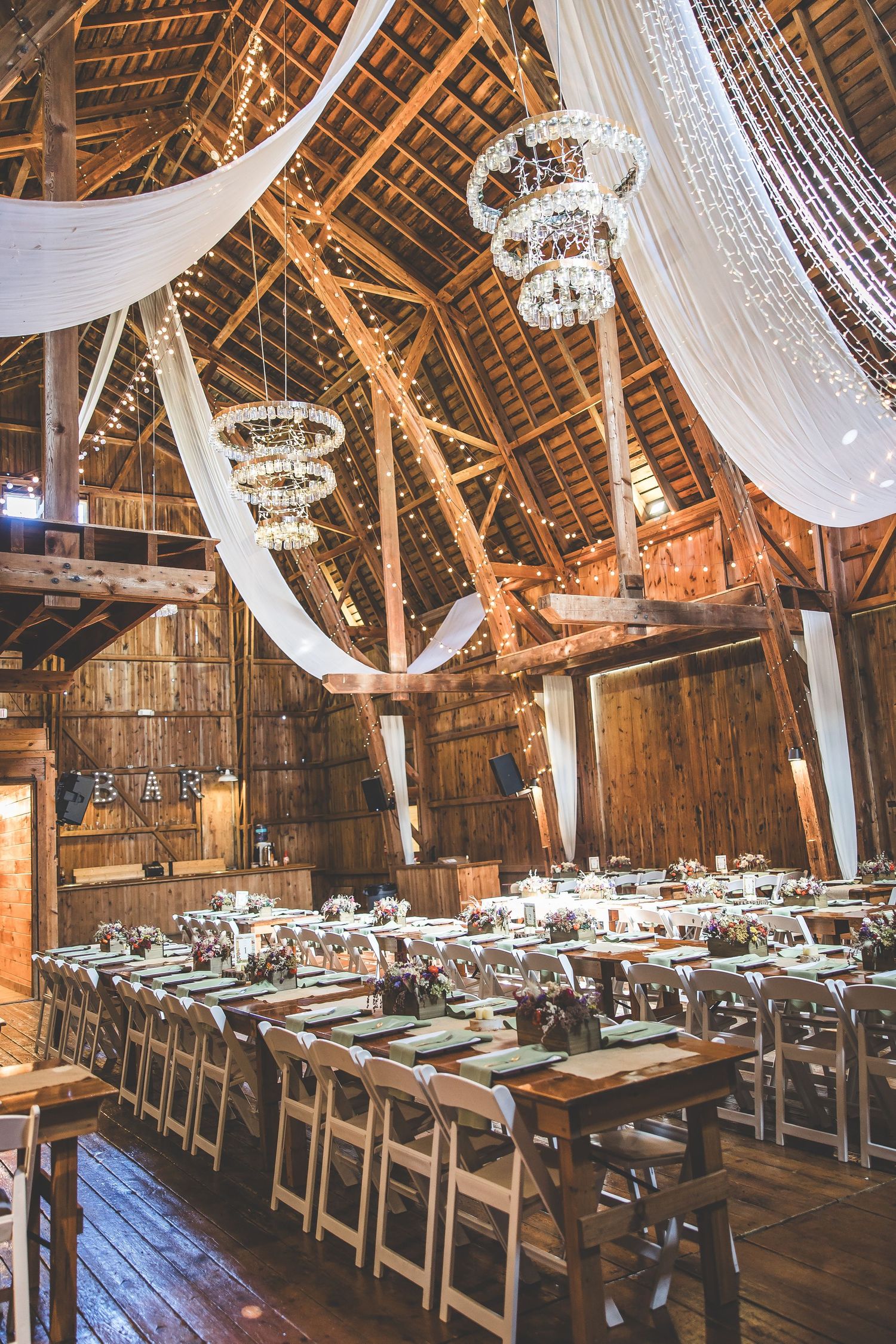 Rustic barn interior decorated with white draping, chandeliers and long wooden tables for wedding reception.