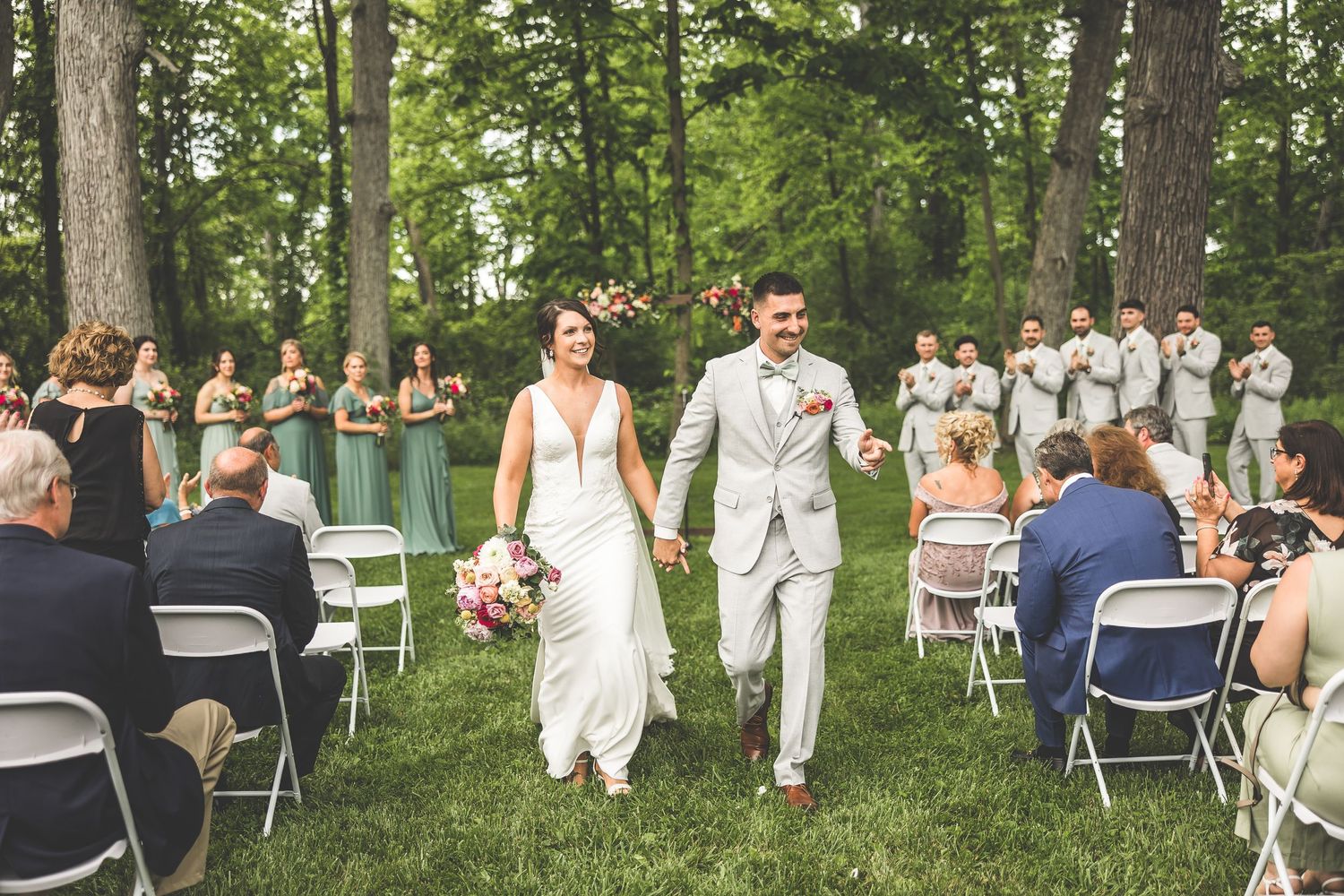 Outdoor wedding ceremony with guests seated on white chairs surrounded by tall trees.