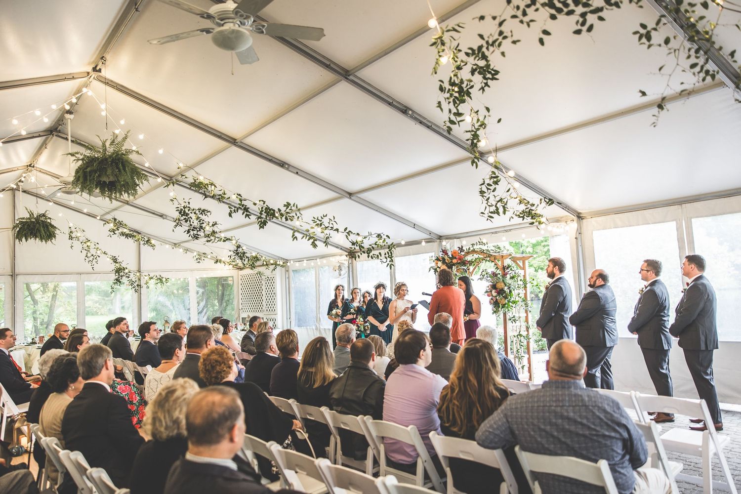 Wide view of wedding ceremony in white tent with guests seated and string lights overhead.