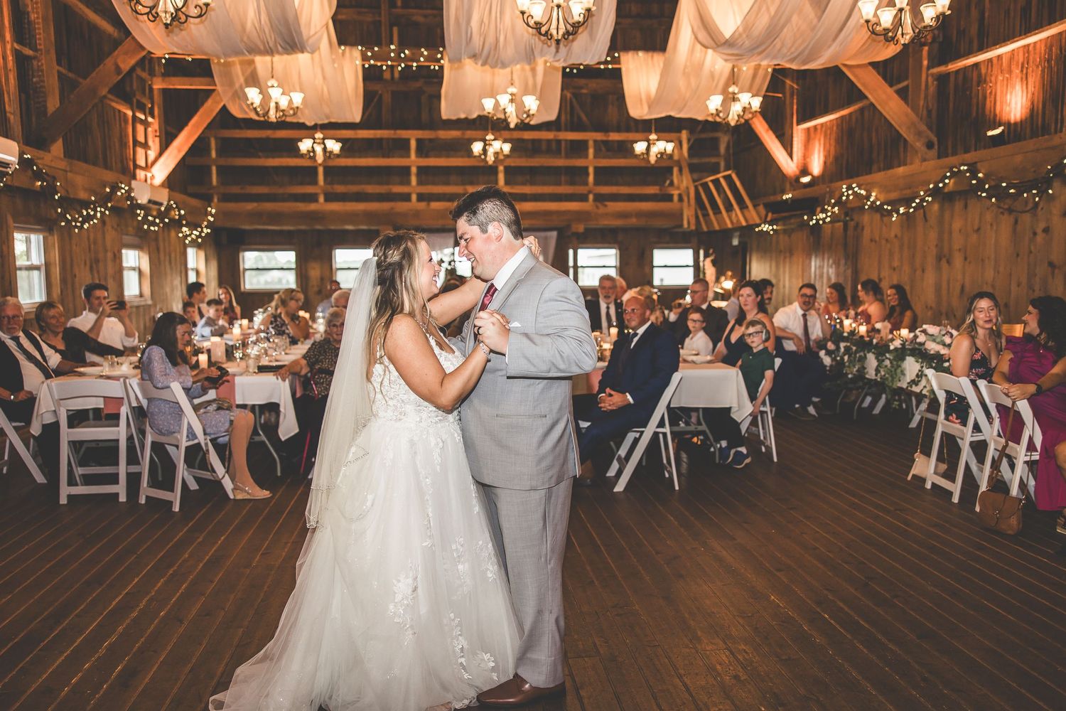 Newlyweds dance surrounded by guests in a beautifully decorated barn with chandeliers and draping.