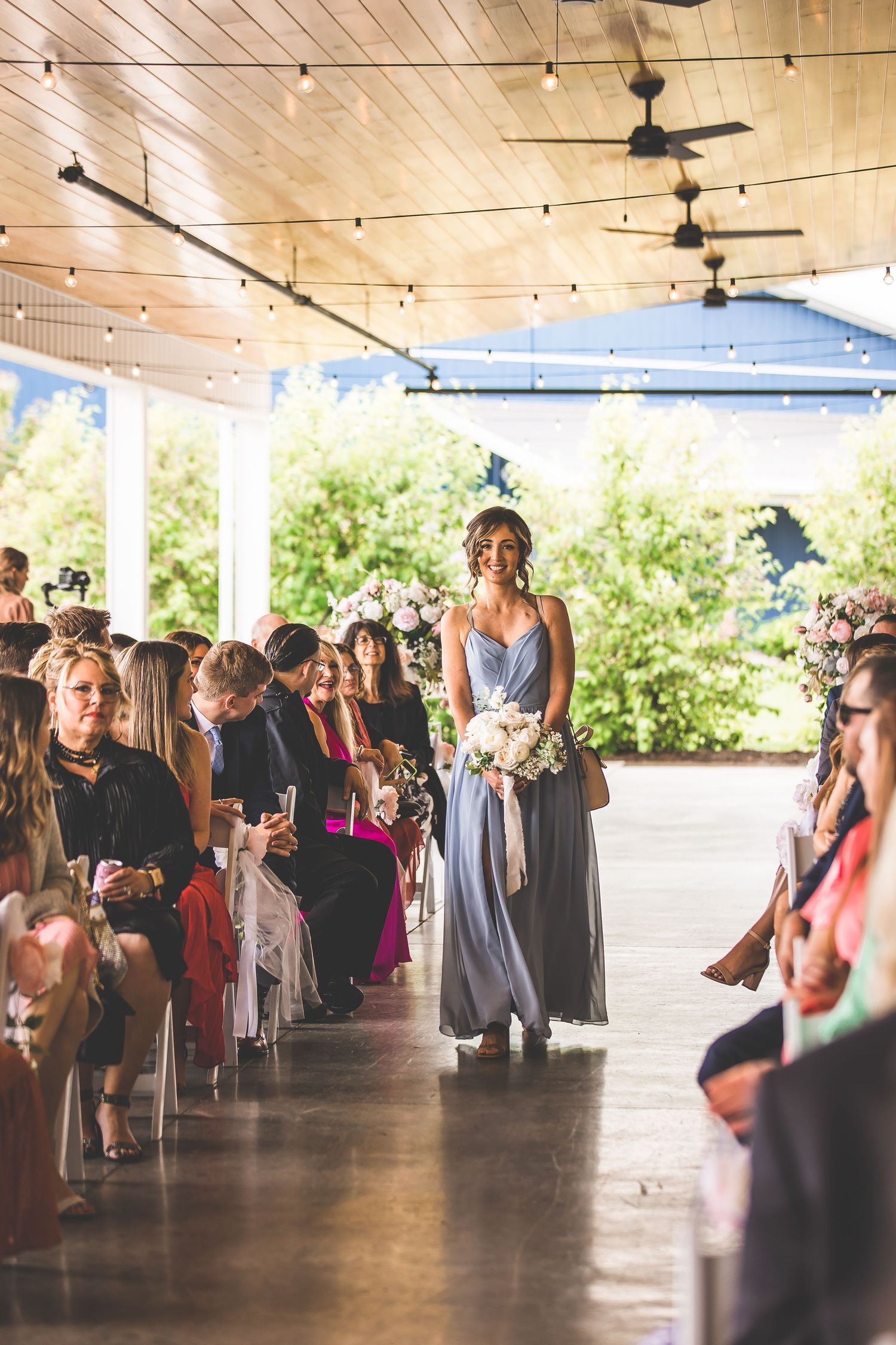 Wedding guests seated in rows at an outdoor covered venue with wooden ceiling and string lights.