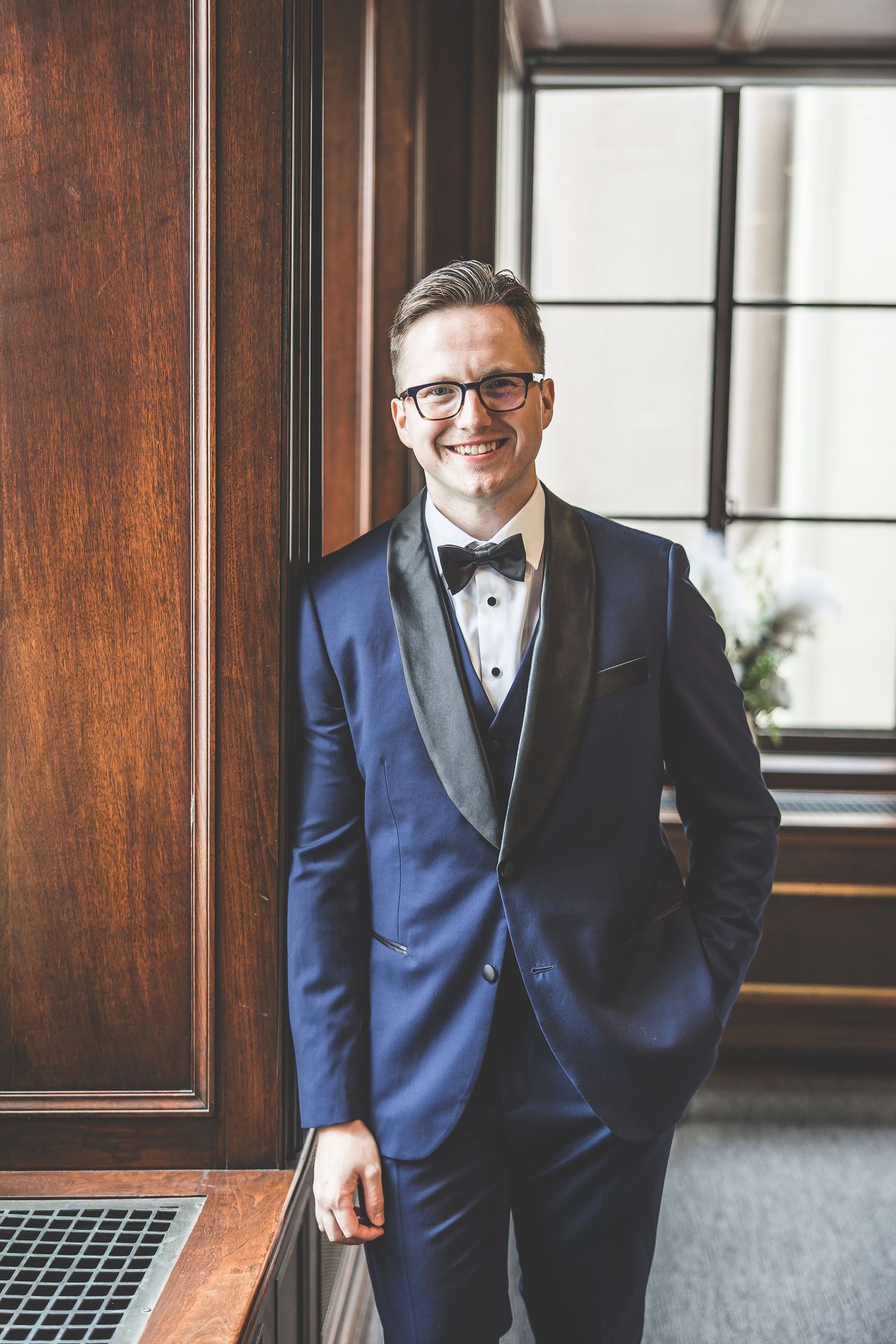 Person in navy tuxedo and bowtie poses confidently by window in formal setting.