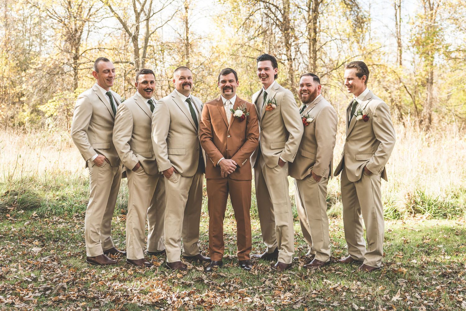 Groomsmen in beige suits pose with the groom in brown for a formal wedding photo outdoors.