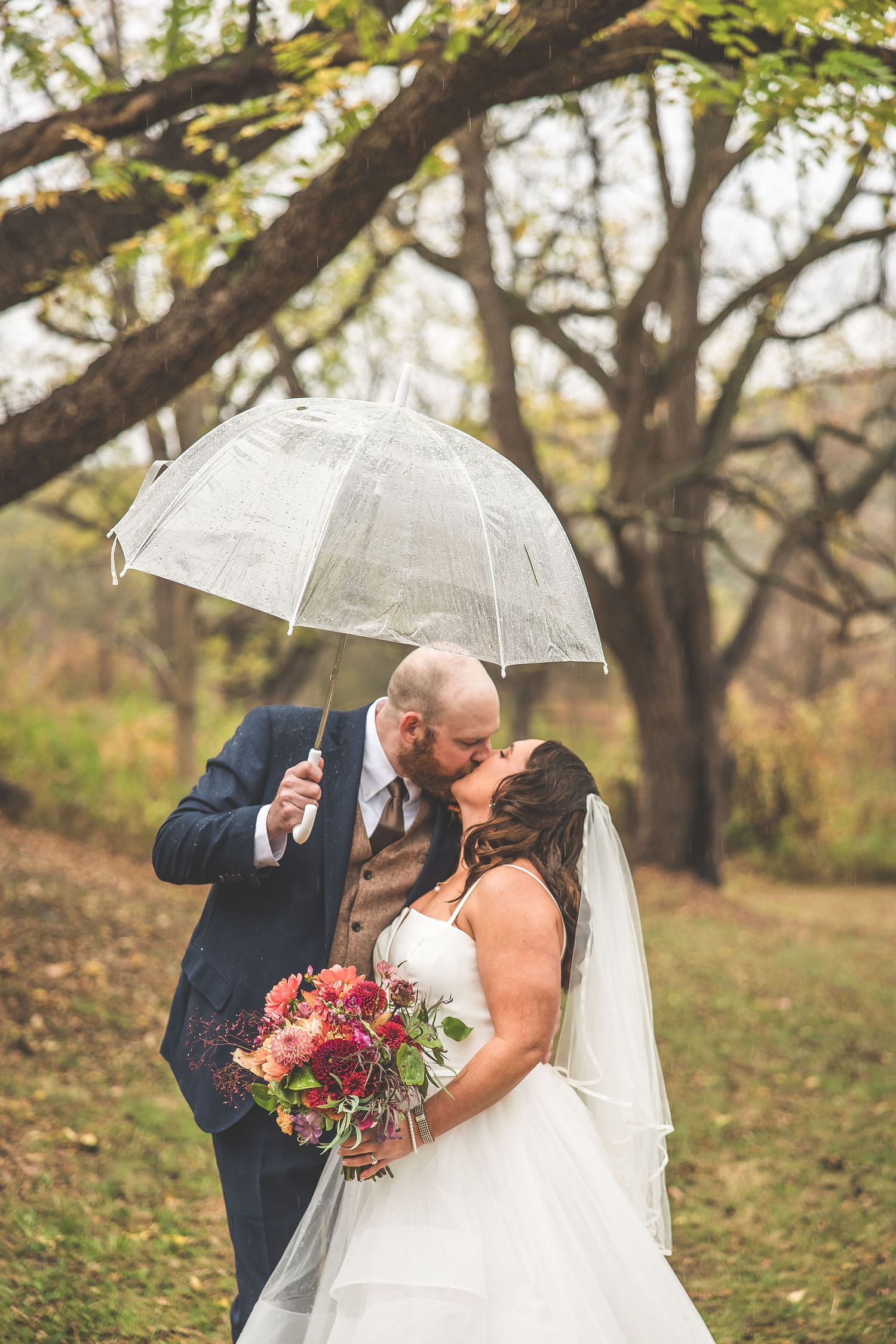 Wedding couple shares romantic moments under clear umbrellas in a wooded autumn setting.
