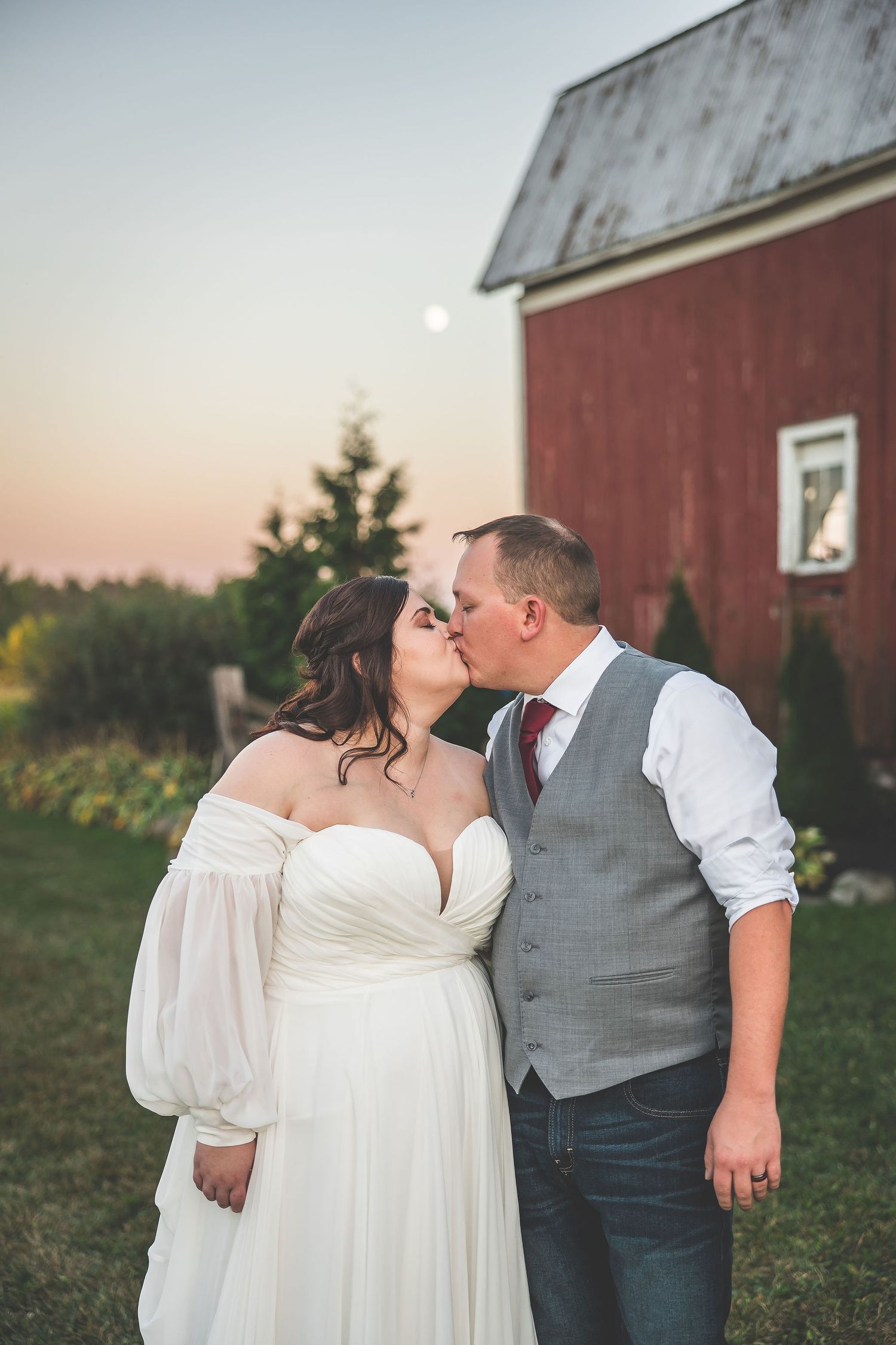 A romantic wedding couple shares a tender moment outside a rustic red barn at sunset.
