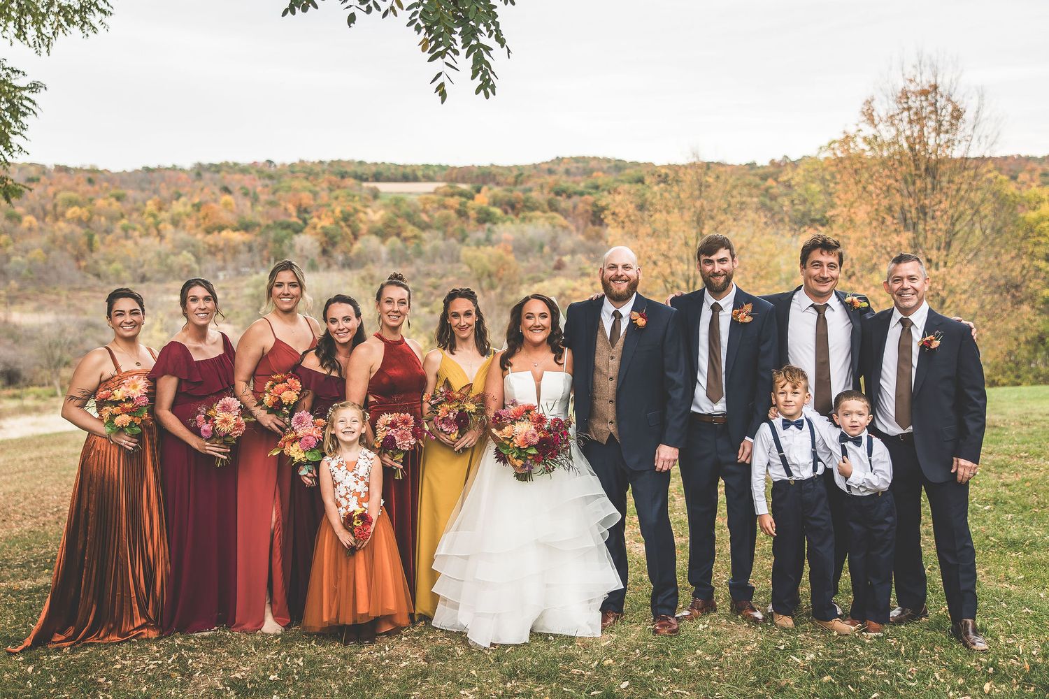 Full wedding party in burgundy dresses and navy suits poses together on autumn hillside.
