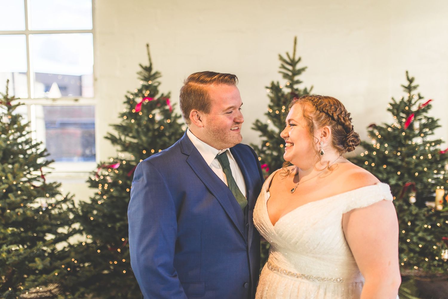 Intimate moment between couple in wedding attire with Christmas trees in background.