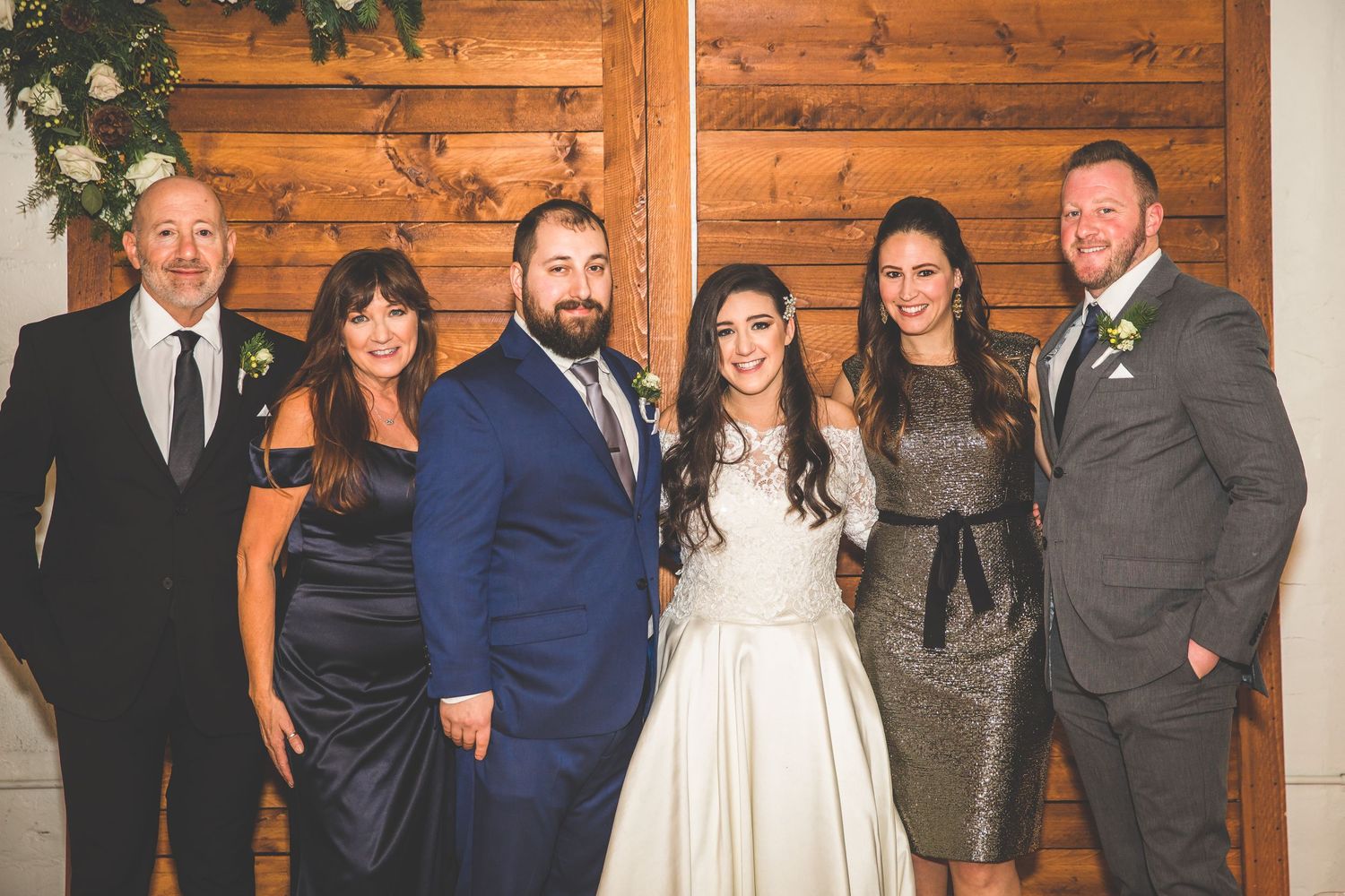 Wedding party of six poses in formal attire against wooden wall with floral decorations.