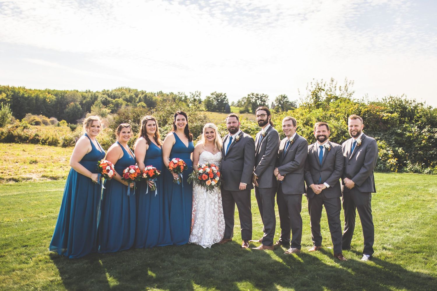 Full wedding party poses on grassy hillside with trees in background.