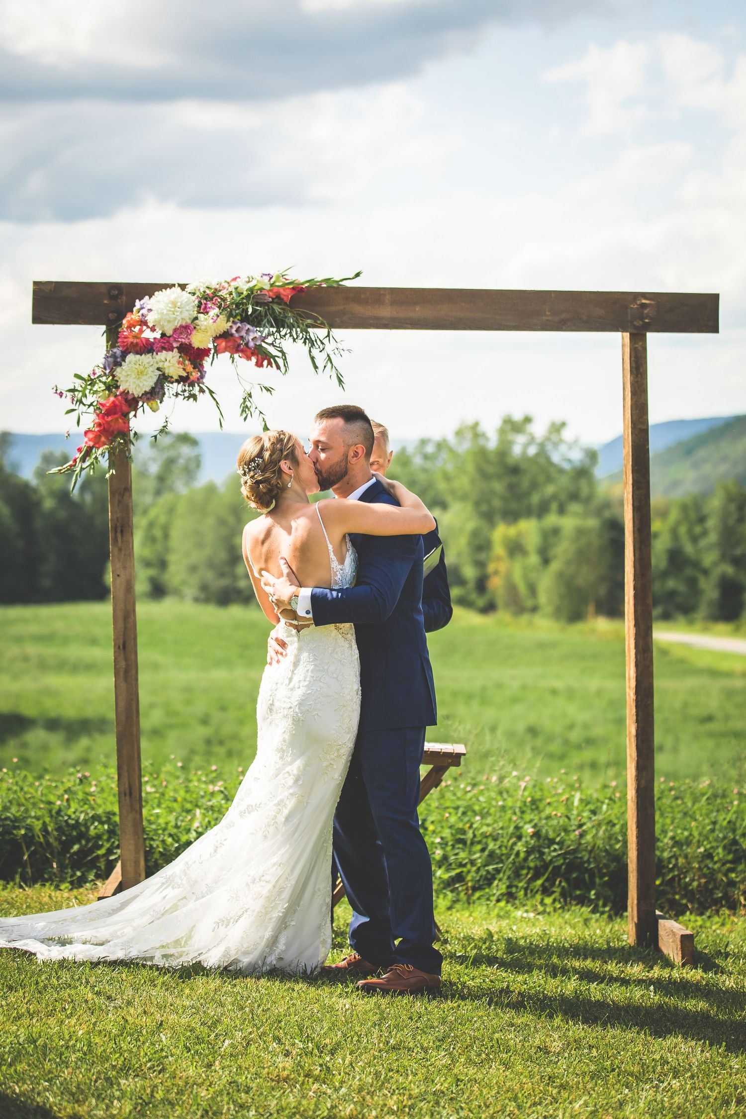 First kiss as newlyweds under wooden ceremony arch decorated with pink flowers on hillside.