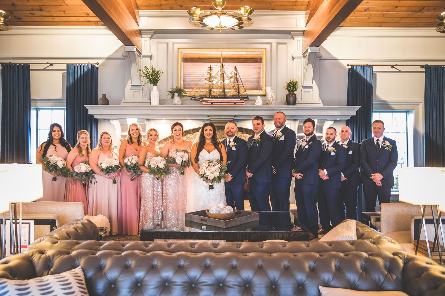 Full wedding party poses together in elegant venue with wooden beams and chandelier lighting.