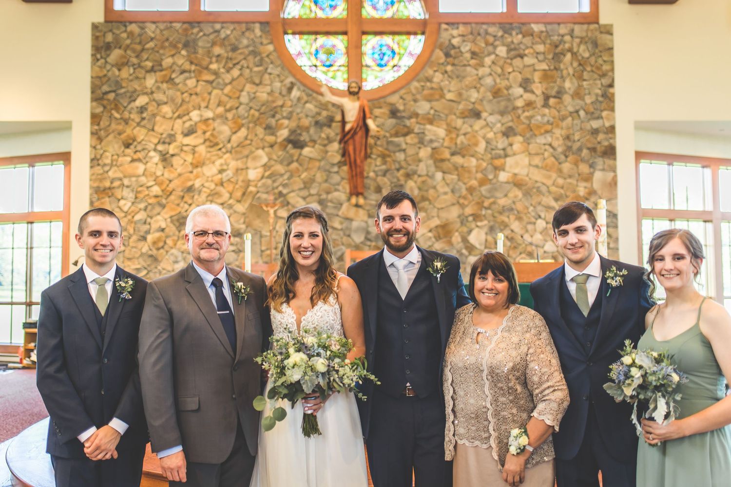Wedding group portrait with bridesmaids in sage green dresses and groomsmen in dark suits.