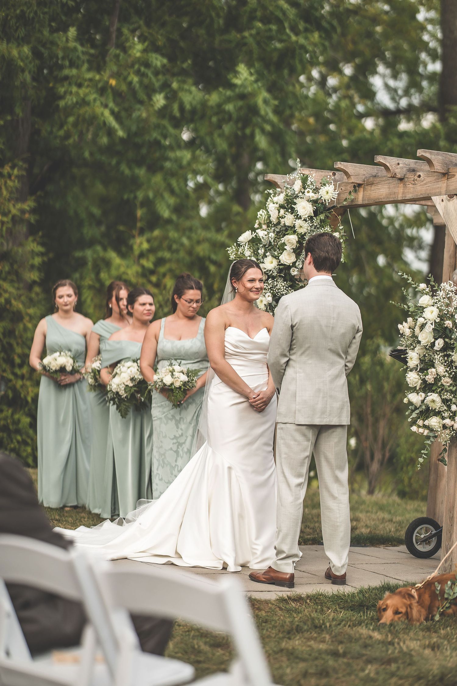 Wedding party in sage green and beige attire poses during outdoor ceremony.
