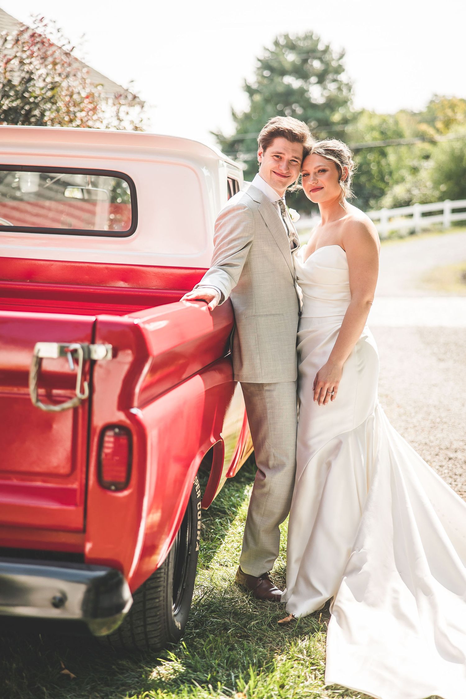 Wedding couple poses beside vintage red Chevrolet truck in outdoor countryside setting.