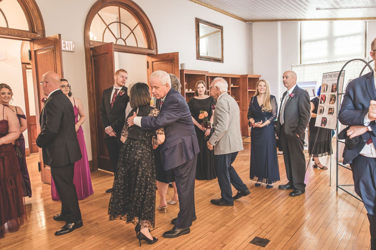 Wedding guests mingle in an elegant reception hall with wooden floors and arched windows.