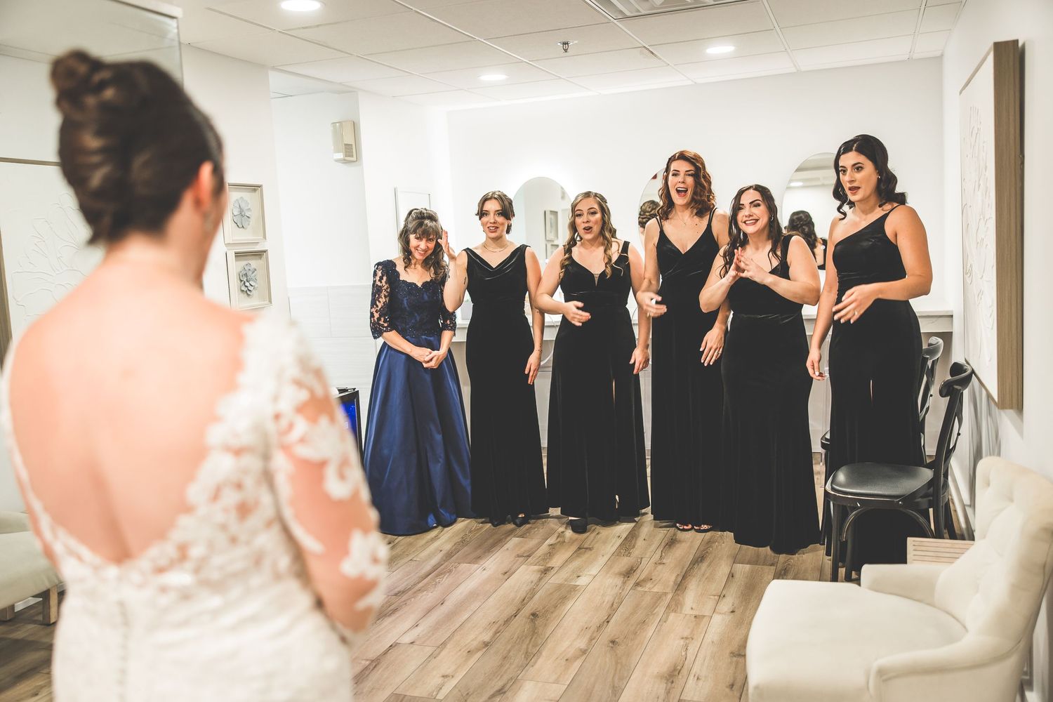 Bridesmaids in black dresses line up along a white wall during wedding preparations.