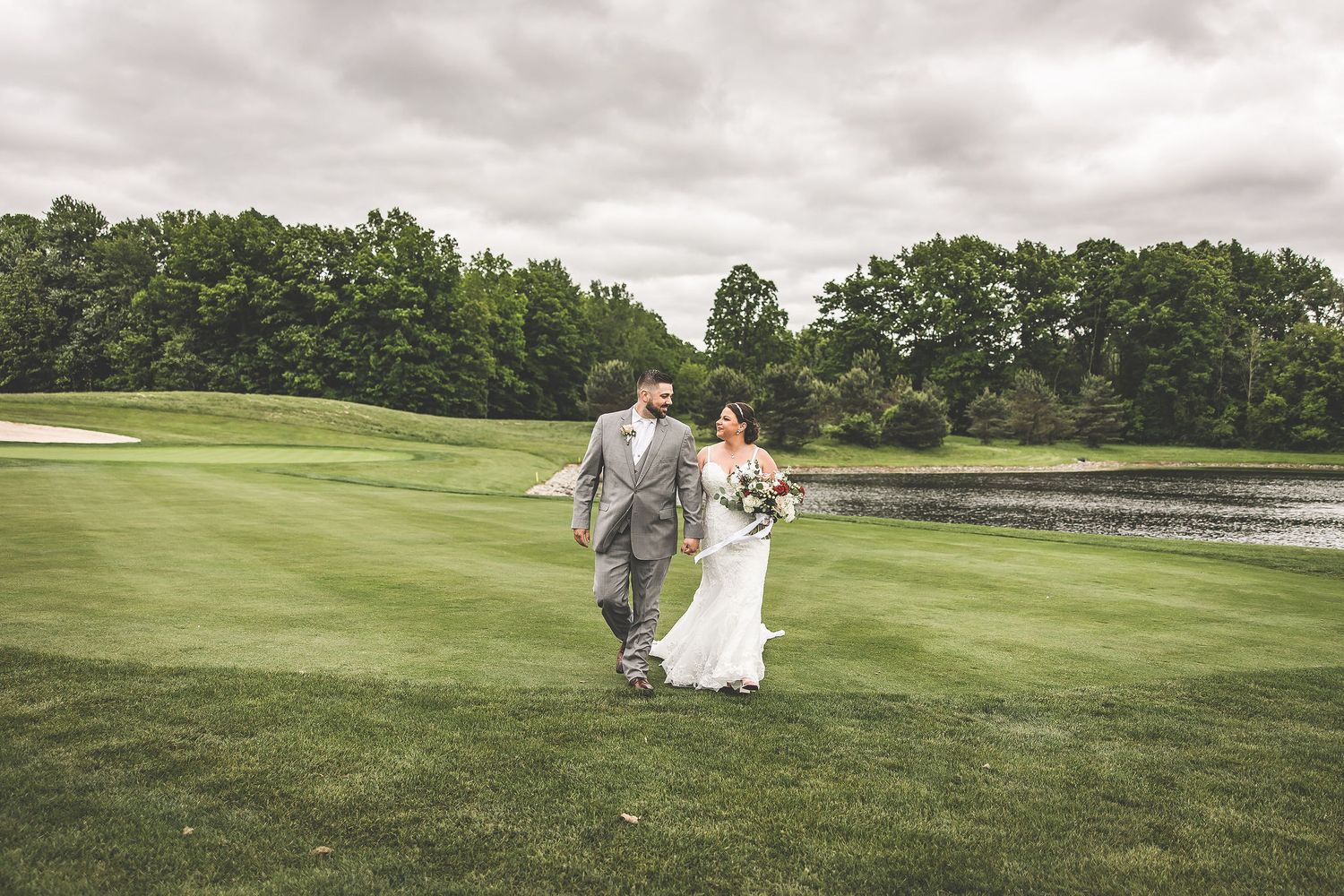 A couple walks across a lush golf course on their wedding day with overcast skies and a pond in the background.