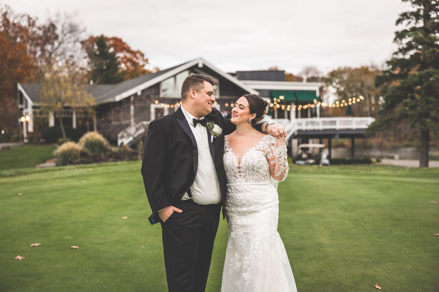 Wedding couple embraces on a scenic lawn with autumn trees and buildings in the background.