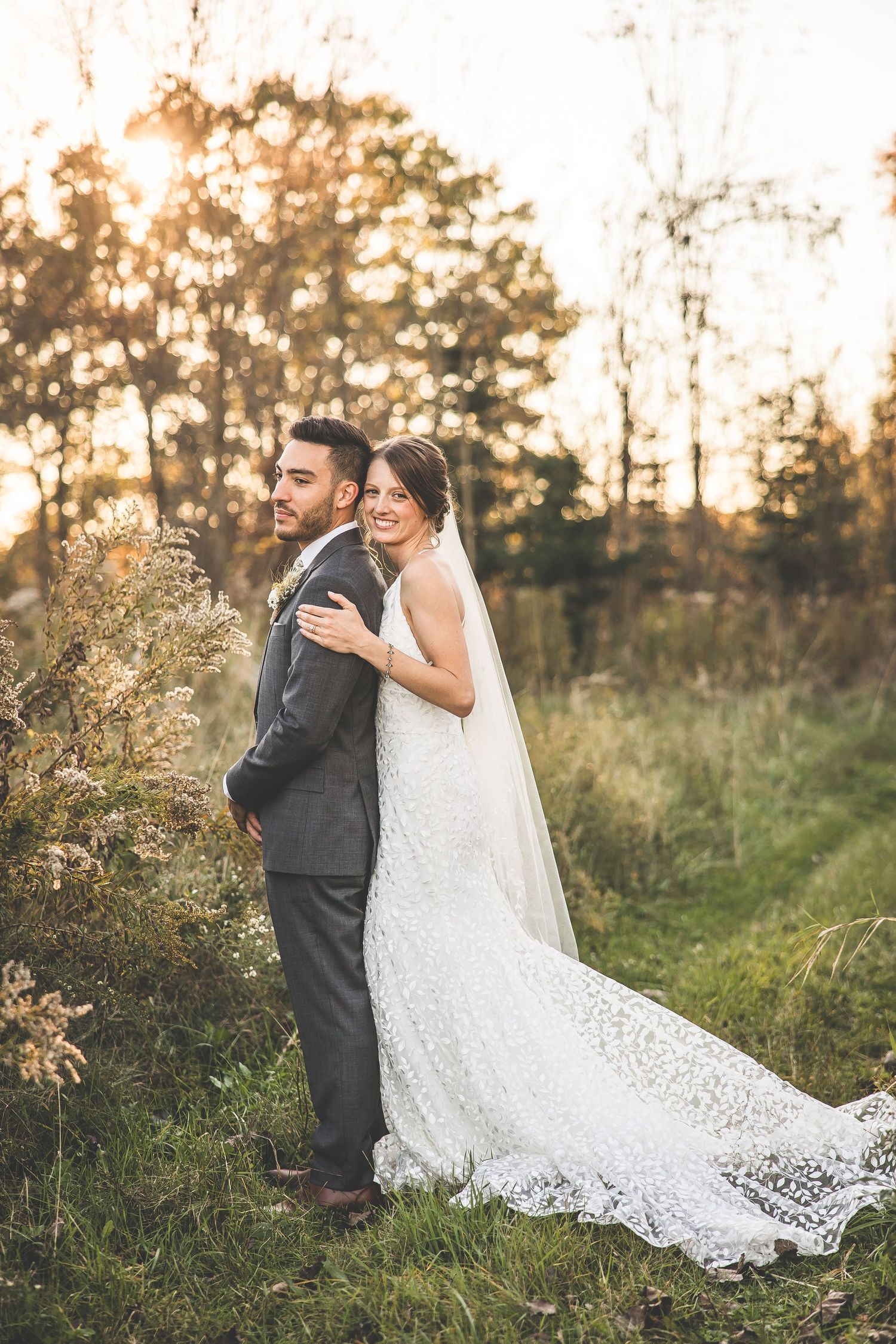 Wedding couple in rustic outdoor setting with long lace dress trailing on autumn grass.