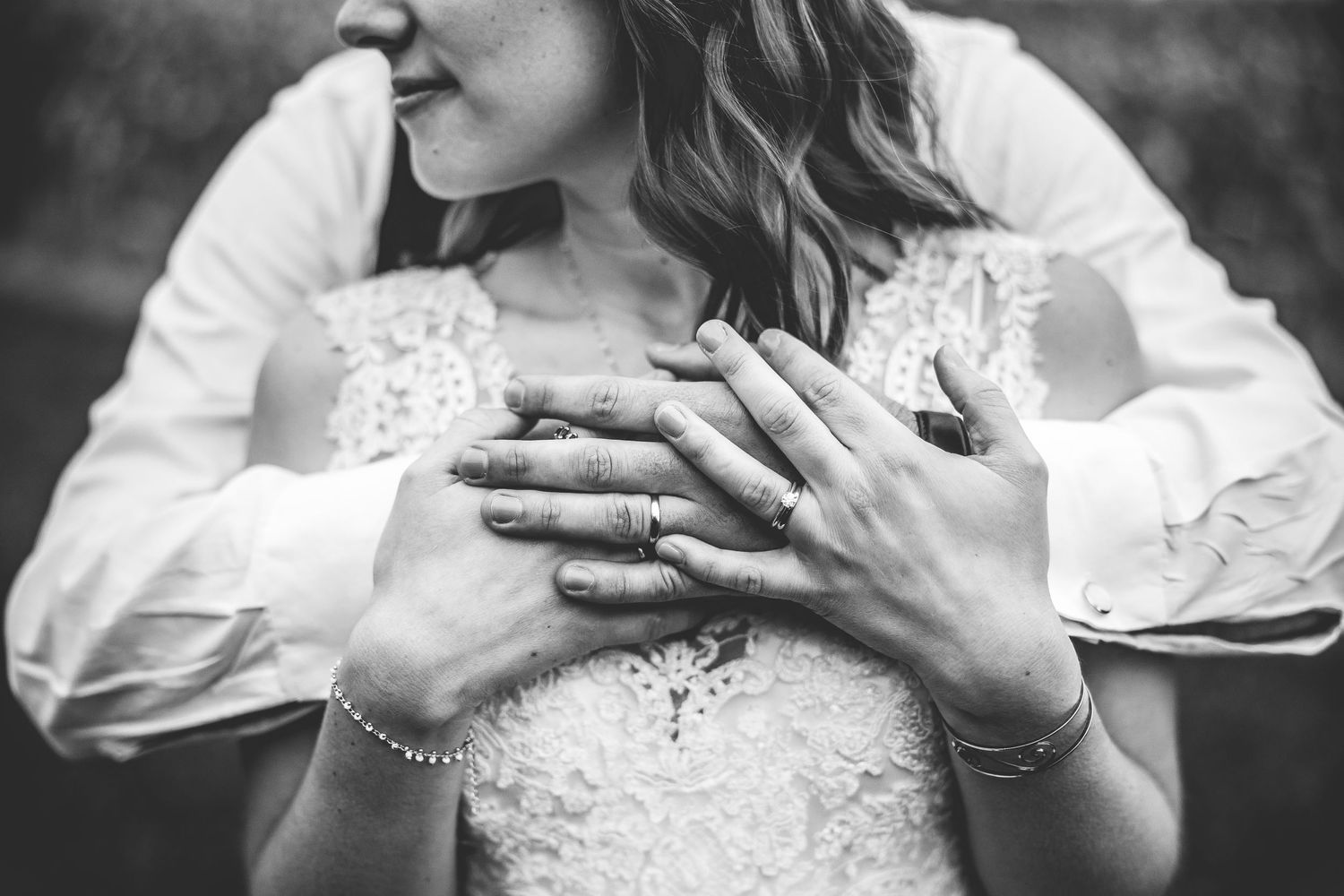 Black and white close-up of bride's hands with wedding ring resting on lace dress bodice.
