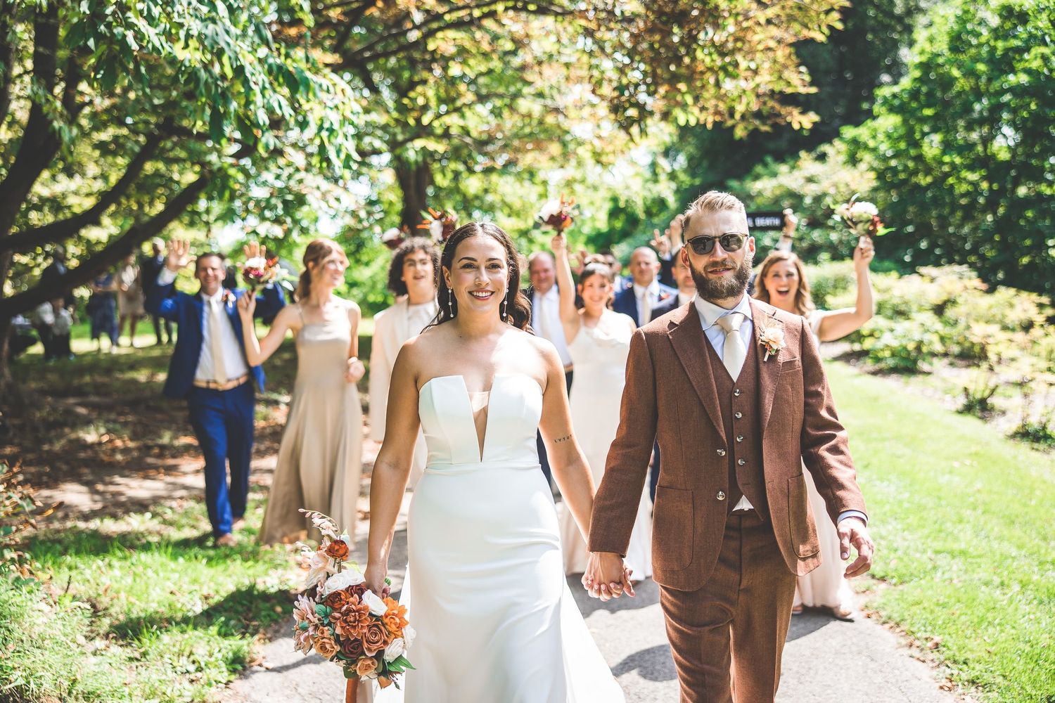 Wedding party walks through garden path with couple leading in brown suit and white dress.