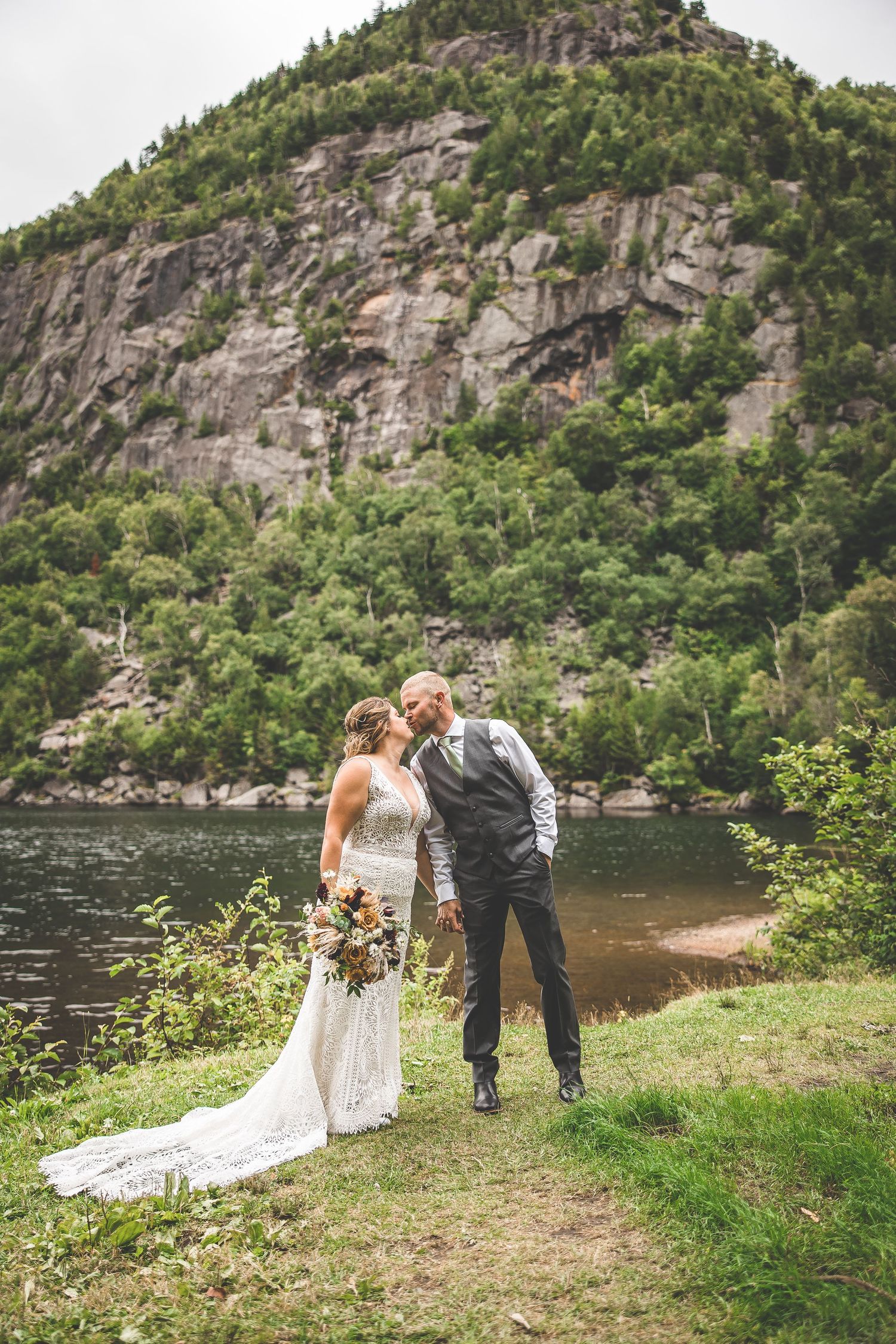 Wedding couple walks along grassy shoreline with dramatic mountain cliffs in background.