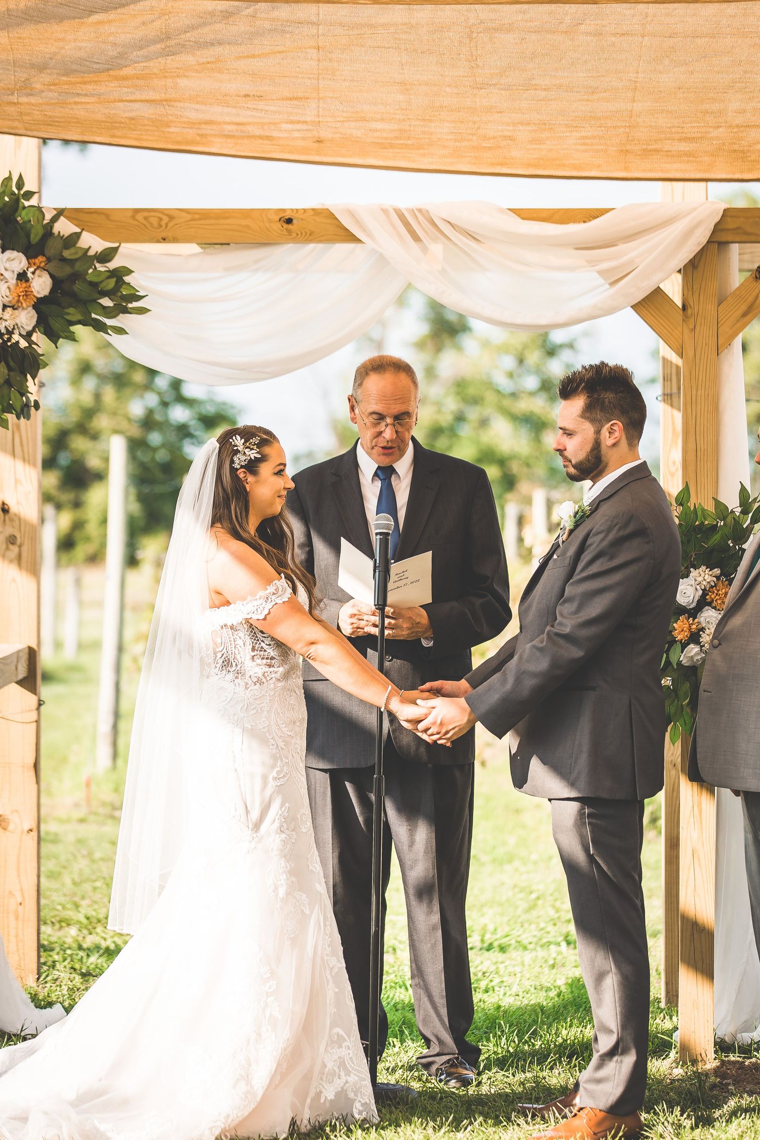 Wedding ceremony taking place under a rustic wooden pavilion with draped white fabric and natural lighting.