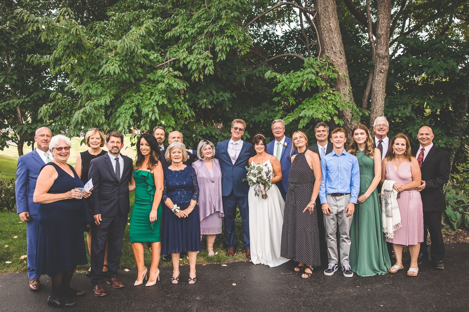 Large group photo of wedding guests gathered under trees in formal attire.