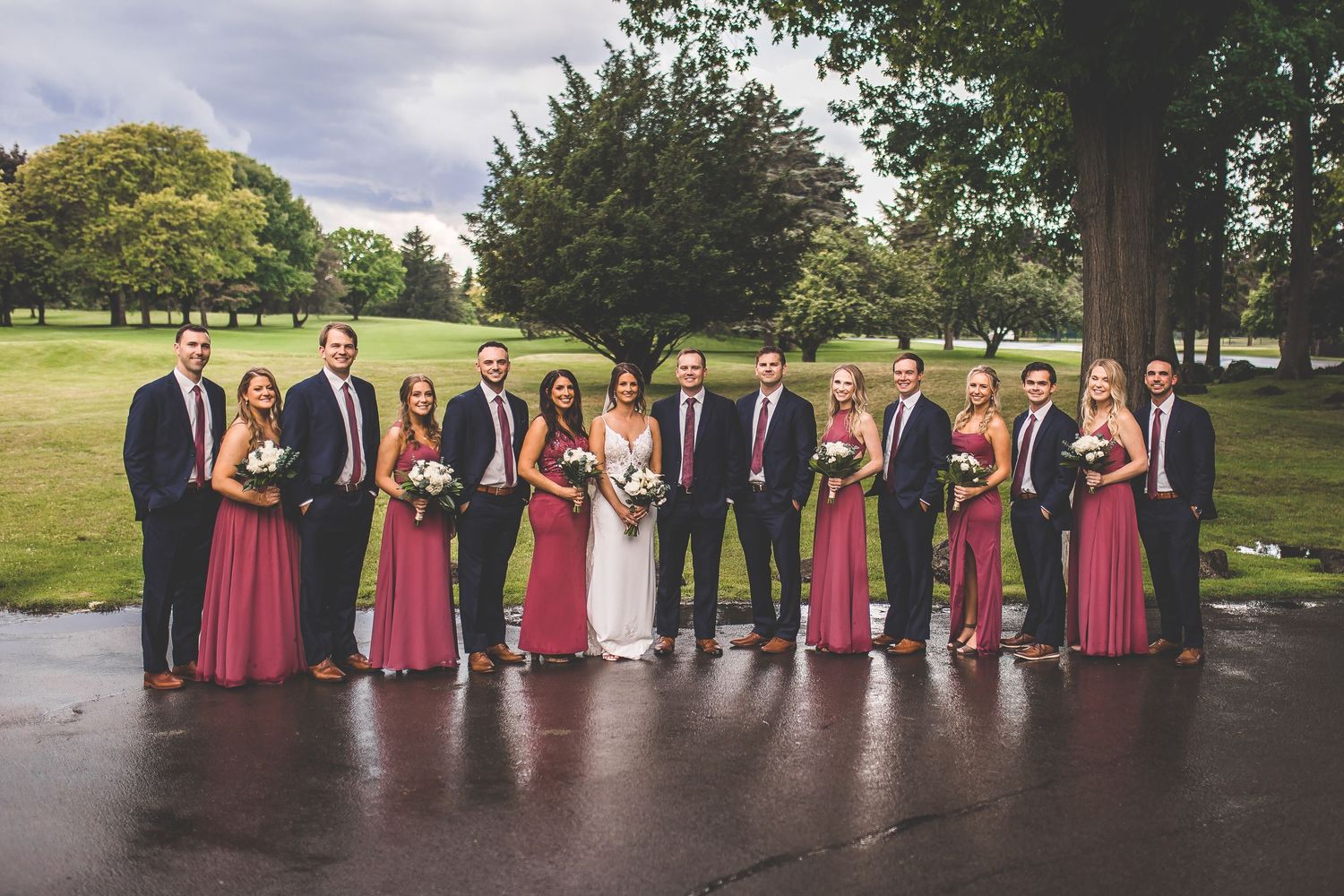 Wedding party poses outdoors in burgundy dresses and navy suits beneath tall trees on a cloudy day.