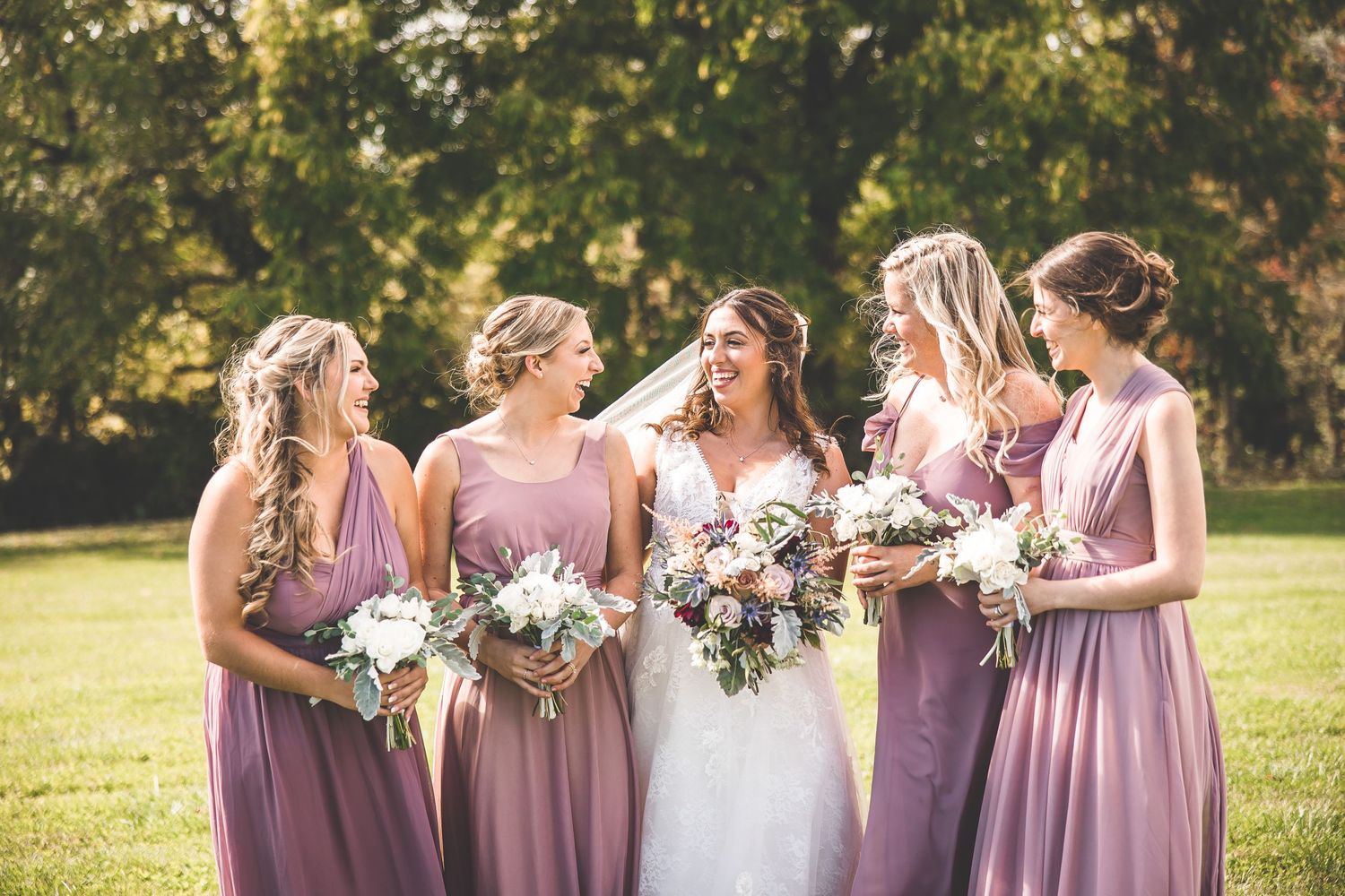 Wedding party shares joyful laughter while holding bouquets in varying shades of pink dresses.