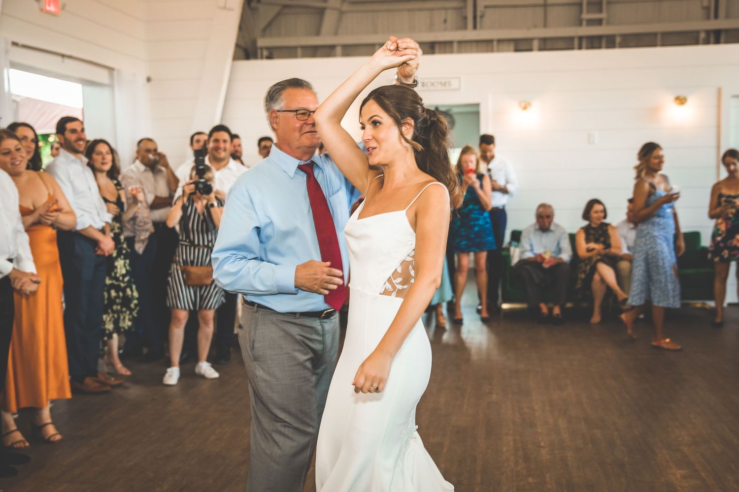 A couple dances together during their wedding reception while guests look on in a bright indoor venue.